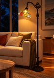 A classic black metal floor lamp with an ornate scrollwork top and a colorful stained-glass shade featuring red, yellow, and green floral motifs, shown lit in a cozy living room next to a beige sofa.