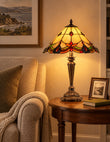 A Tiffany-style table lamp with a stained glass shade and ornate metal base, glowing warmly on a round wooden side table next to a beige armchair in a cozy living room setting.